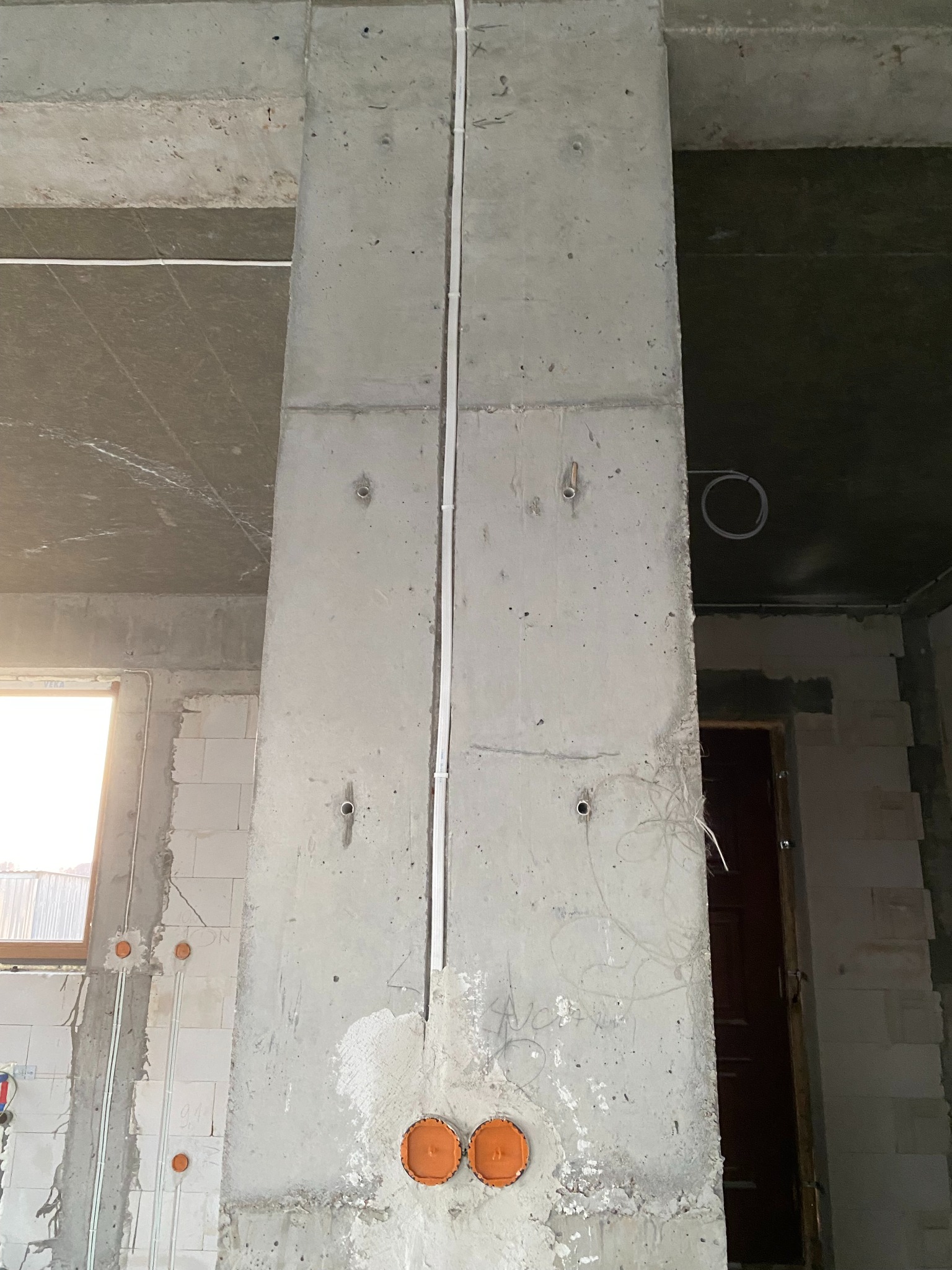 Surowy betonowy filar z poprowadzonymi białymi przewodami elektrycznymi i pomarańczowymi puszkami podtynkowymi w trakcie prac instalacyjnych w nowym budynku.