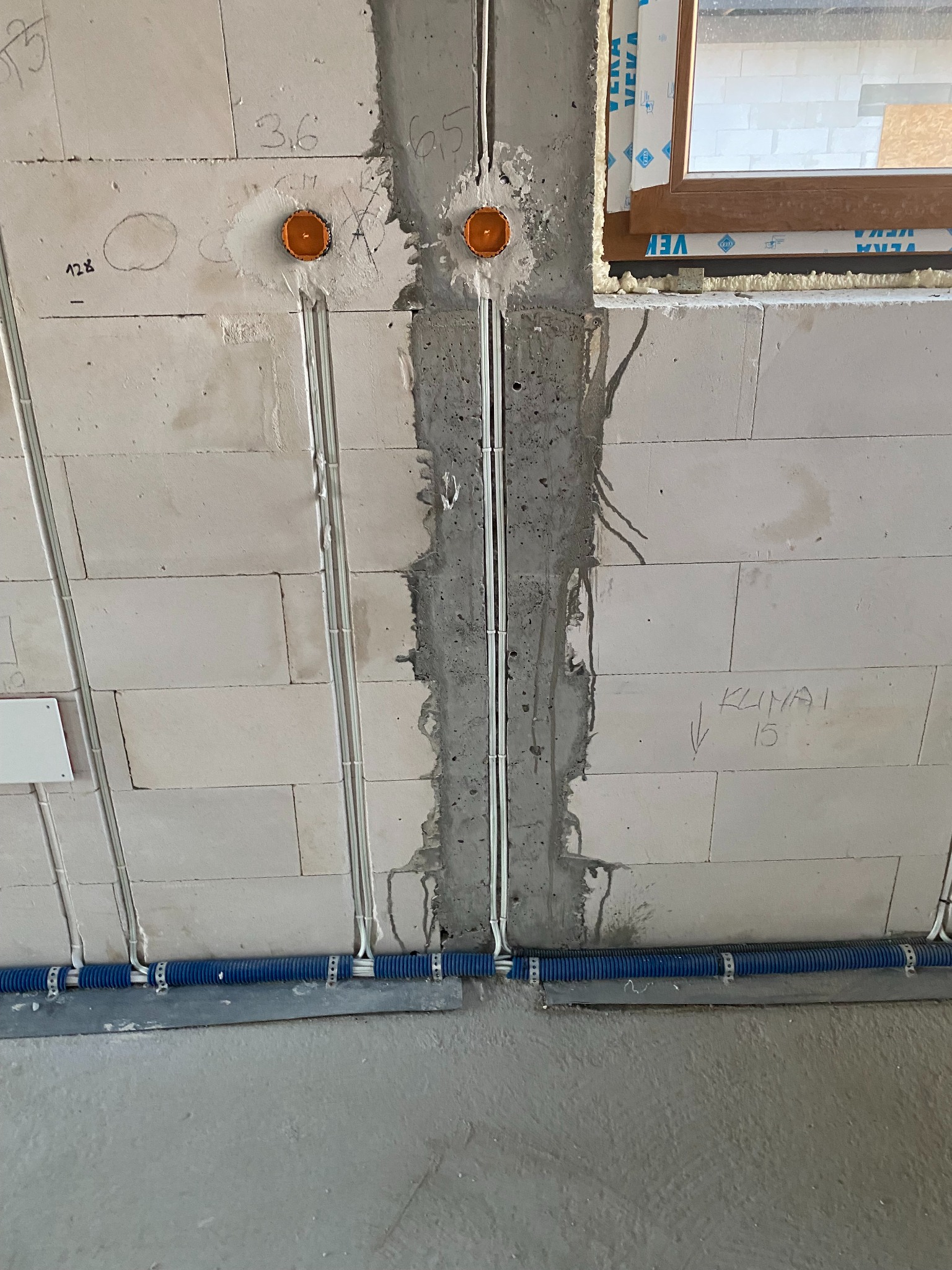 Instalacja elektryczna w surowym stanie: kable w peszlu, puszki instalacyjne w ścianie z betonu komórkowego z widocznymi oznaczeniami, okno w tle.