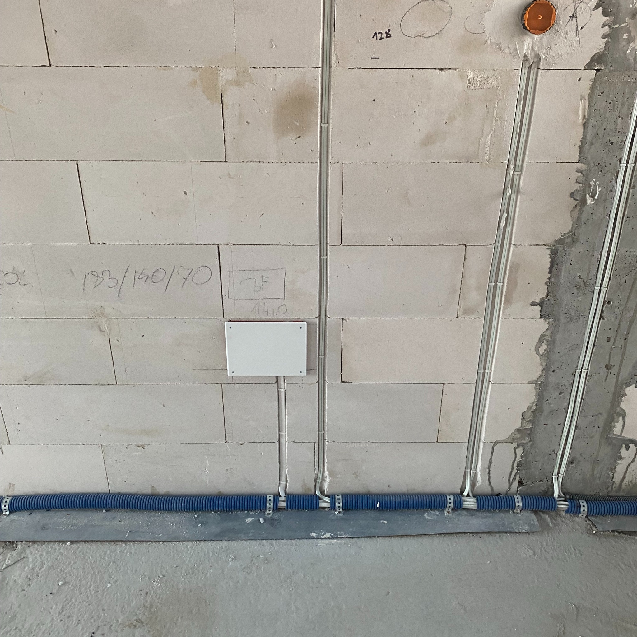 Ściana z betonu komórkowego z widocznymi przewodami elektrycznymi w białych rurkach przymocowanych do ściany oraz niebieską rurą karbowaną przy podłodze. Na ścianie widoczne napisy i oznaczenia...