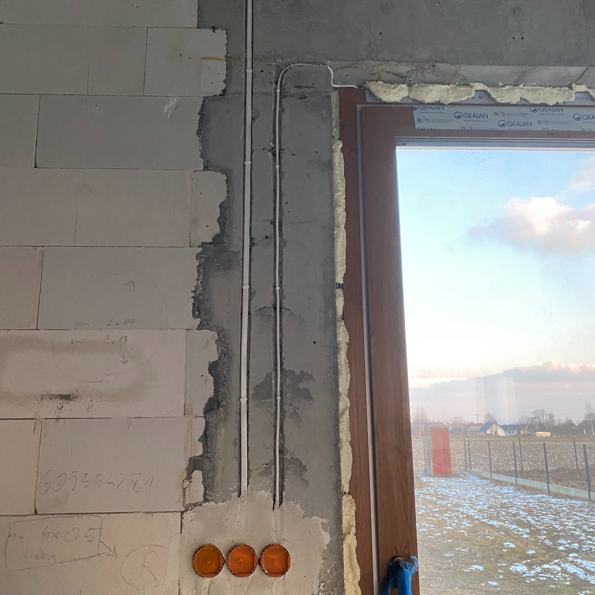 Surowe wnętrze w trakcie wykańczania: widoczne przewody elektryczne prowadzone po ścianie z betonu komórkowego, trzy pomarańczowe puszki podtynkowe i okno z pianką montażową. Na ścianie widoczne...