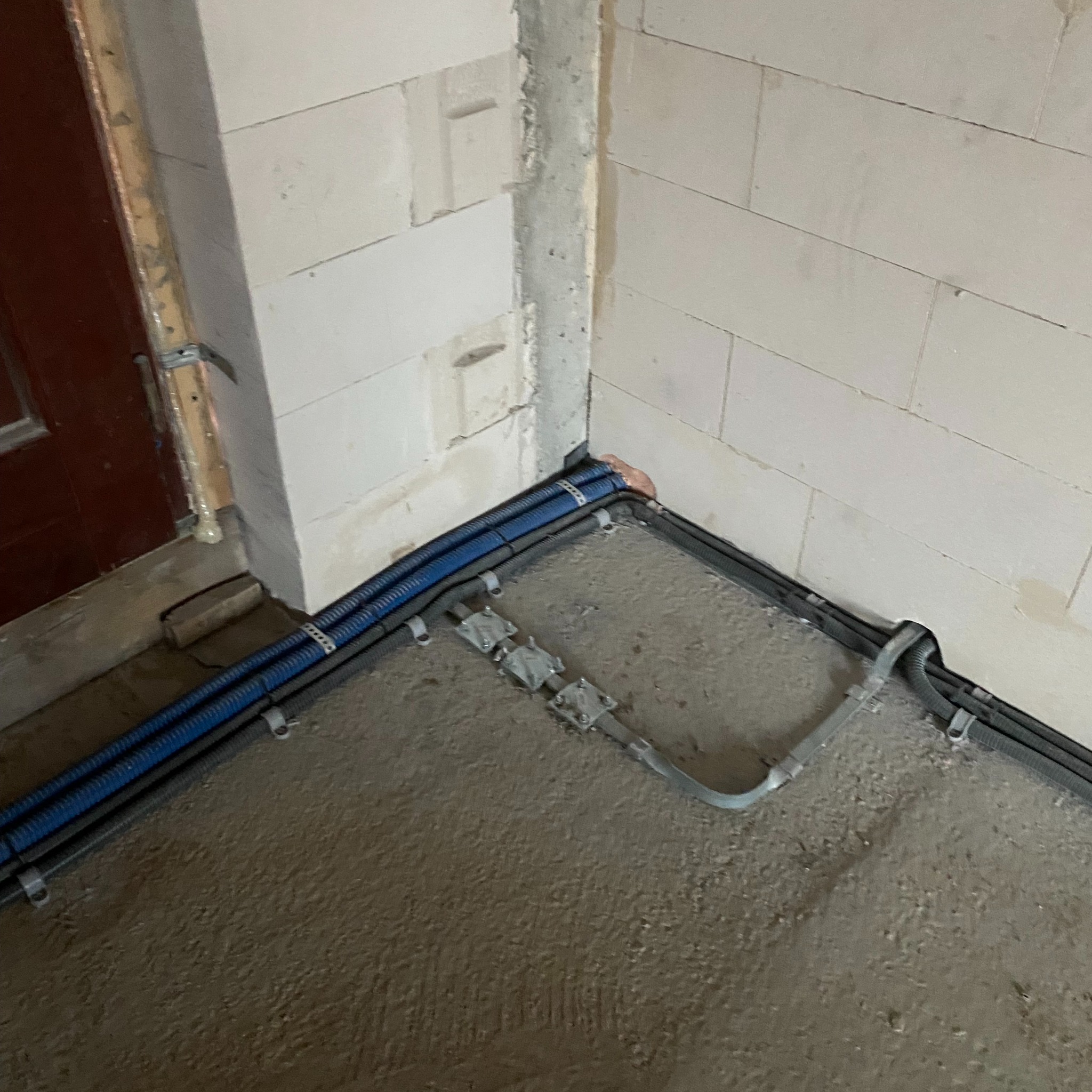 Szczegółowe ujęcie instalacji elektrycznej w korycie, prowadzonej wzdłuż ściany z betonu komórkowego i ramy drzwiowej, z widocznymi uchwytami mocującymi i szarymi rurami ochronnymi w surowym stanie.