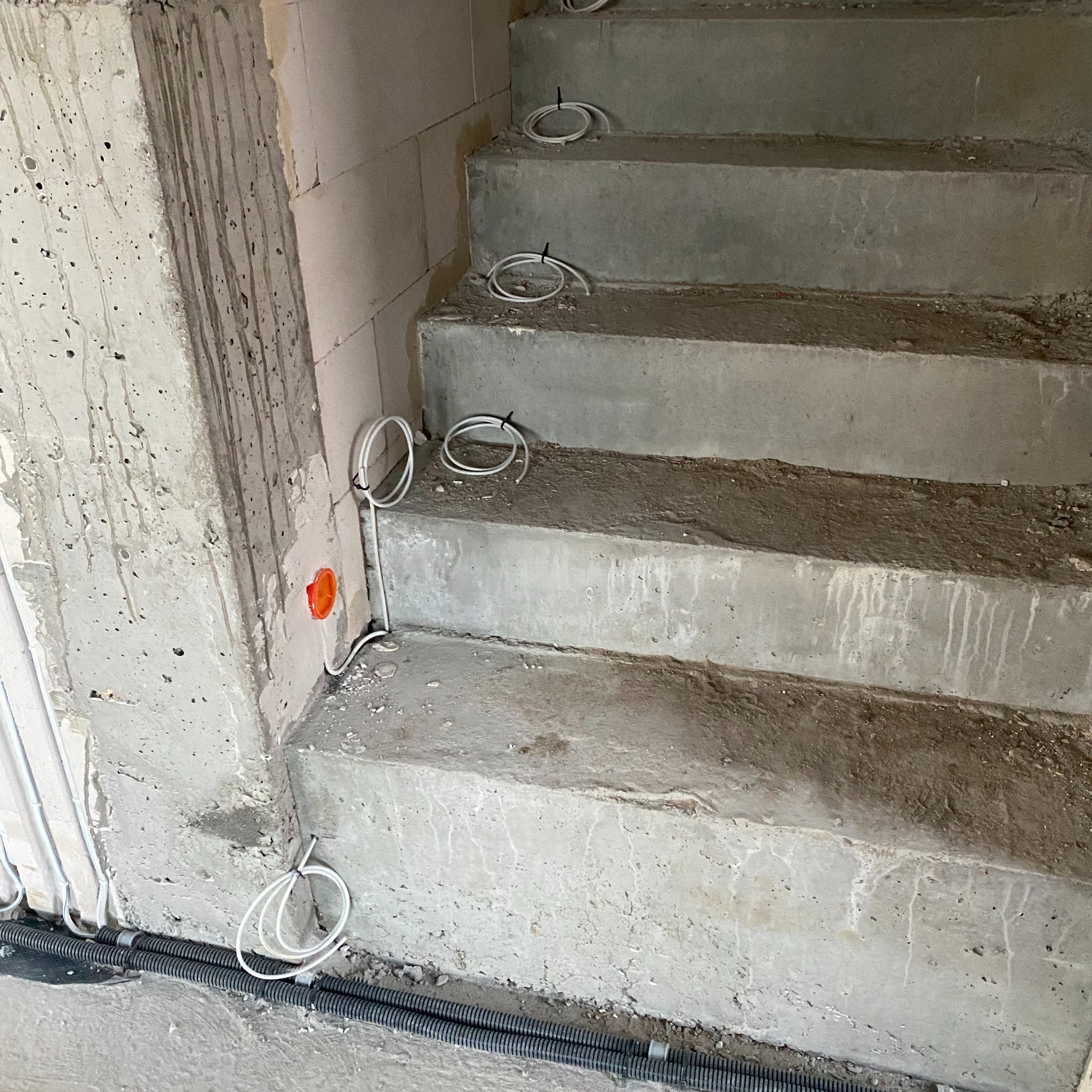 Surowe betonowe schody w trakcie budowy z poprowadzonymi kablami elektrycznymi, widoczne przewody w peszlu przy podłodze i kable zwinięte na stopniach, ściany w stanie surowym.