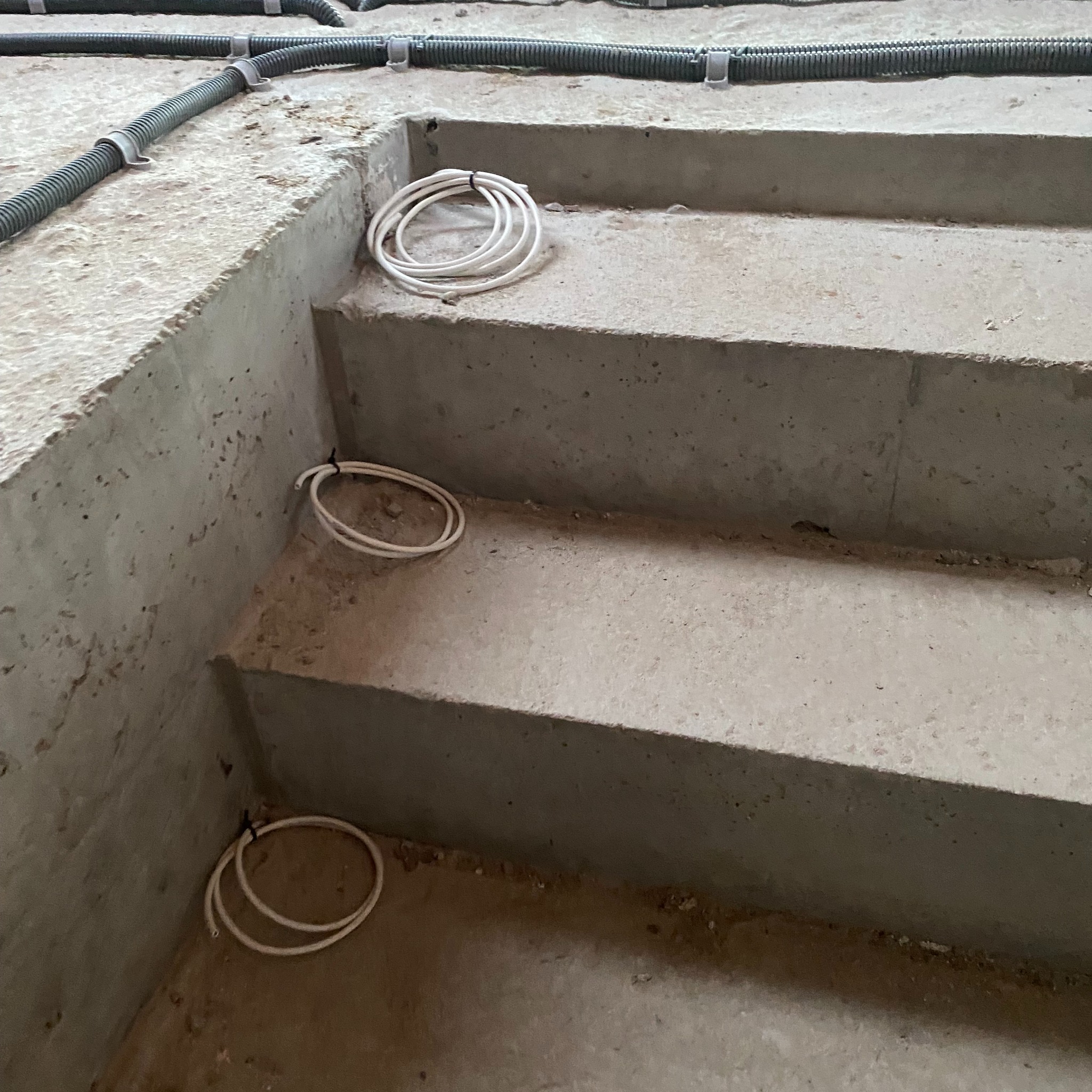 Surowe betonowe schody w trakcie budowy z widocznymi rurami karbowanymi i zwiniętymi przewodami elektrycznymi, zabezpieczonymi opaskami.