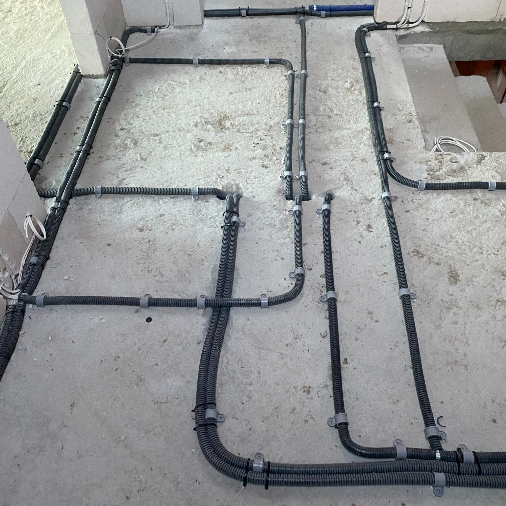 Szare, karbowane rury instalacji elektrycznej prowadzone po surowym, betonowym suficie z widocznymi uchwytami mocującymi i przewodami wystającymi ze ściany.