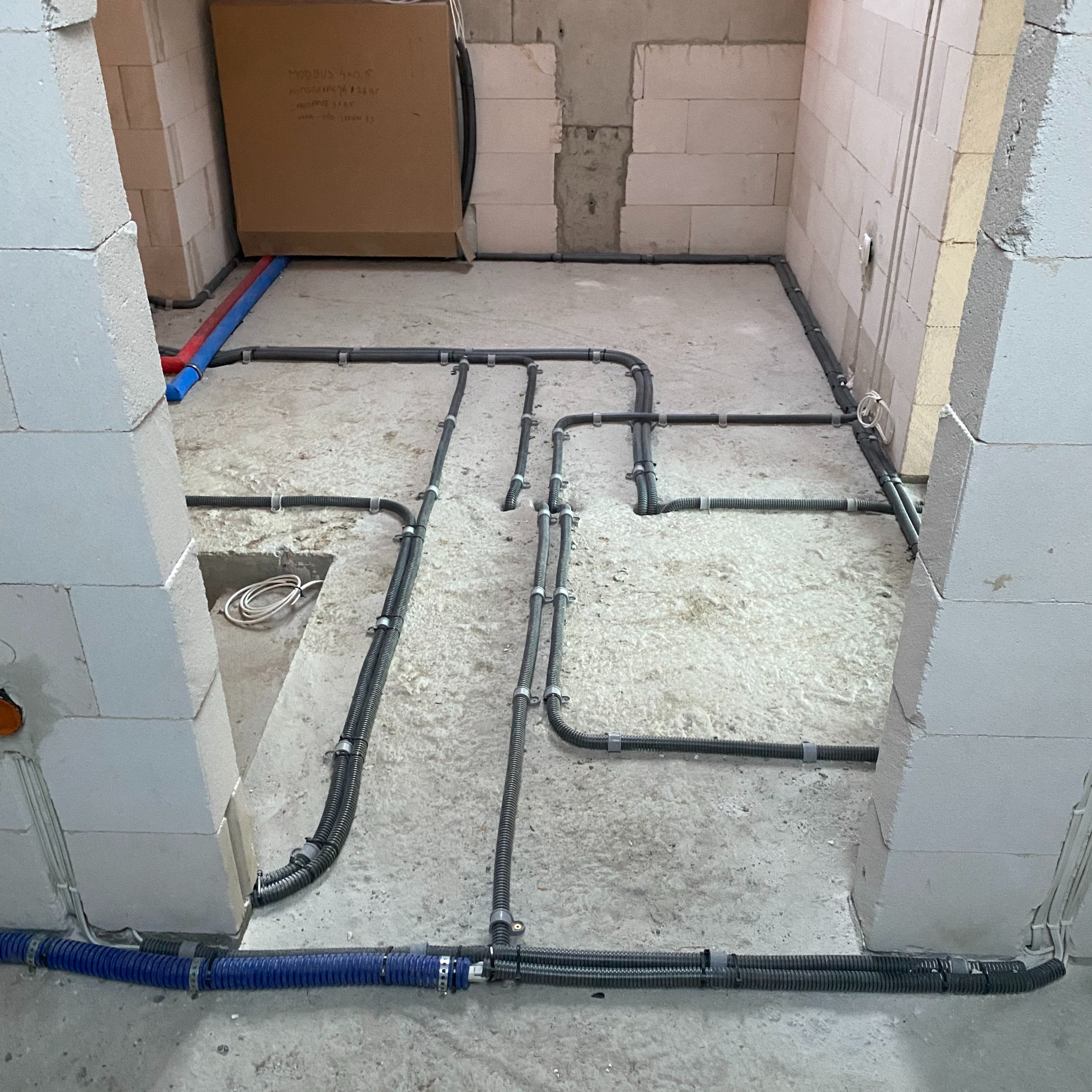 Wnętrze w trakcie prac instalacyjnych: szare rury karbowane prowadzone po podłodze w surowym pomieszczeniu z bloczków, widoczne fragmenty rur niebieskich i czerwonych, kartonowe pudło w rogu.