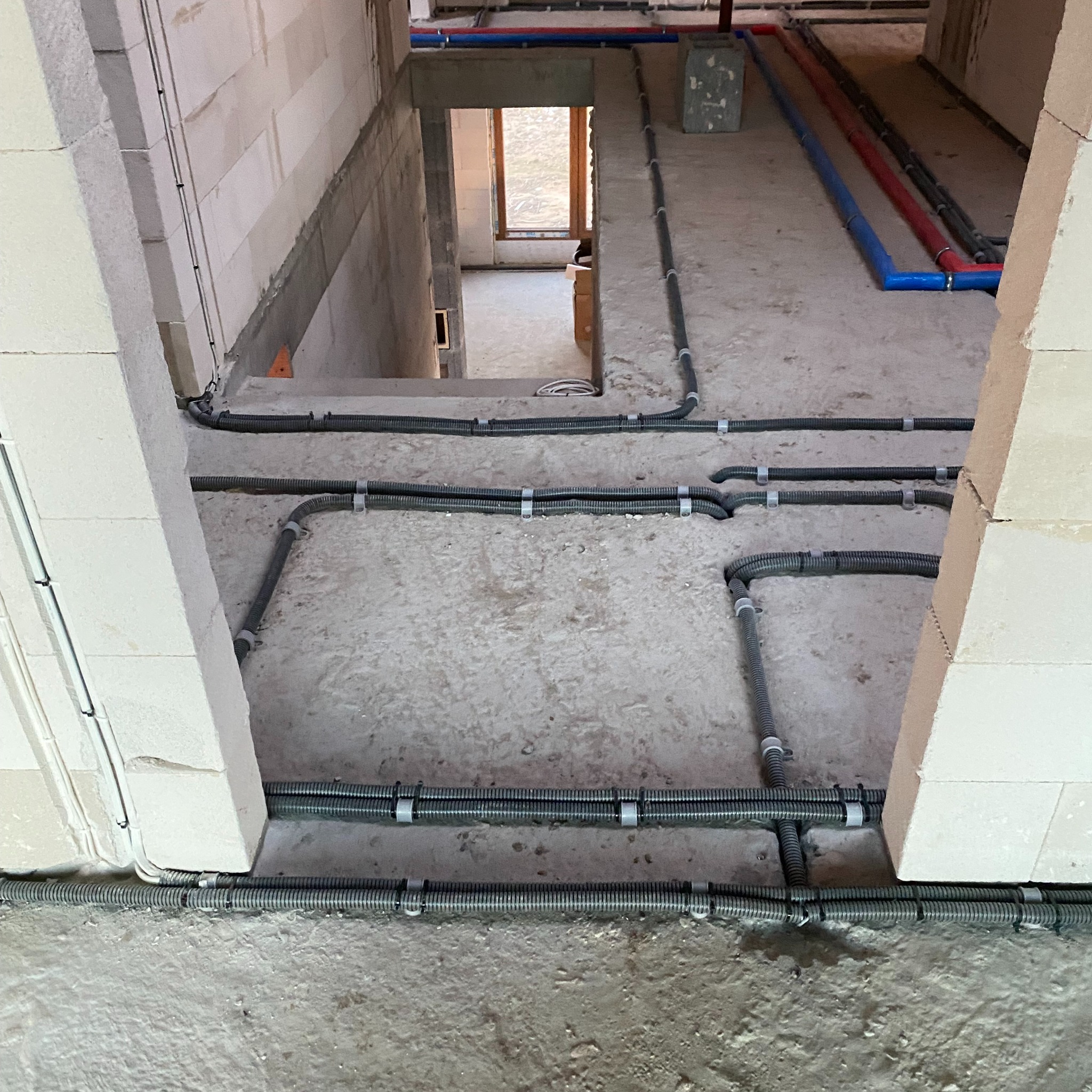 Szare rury karbowane prowadzone wzdłuż podłogi i ścian z betonu komórkowego w surowym stanie, zabezpieczone plastikowymi uchwytami, z widocznymi rurami instalacji wodnej w tle.