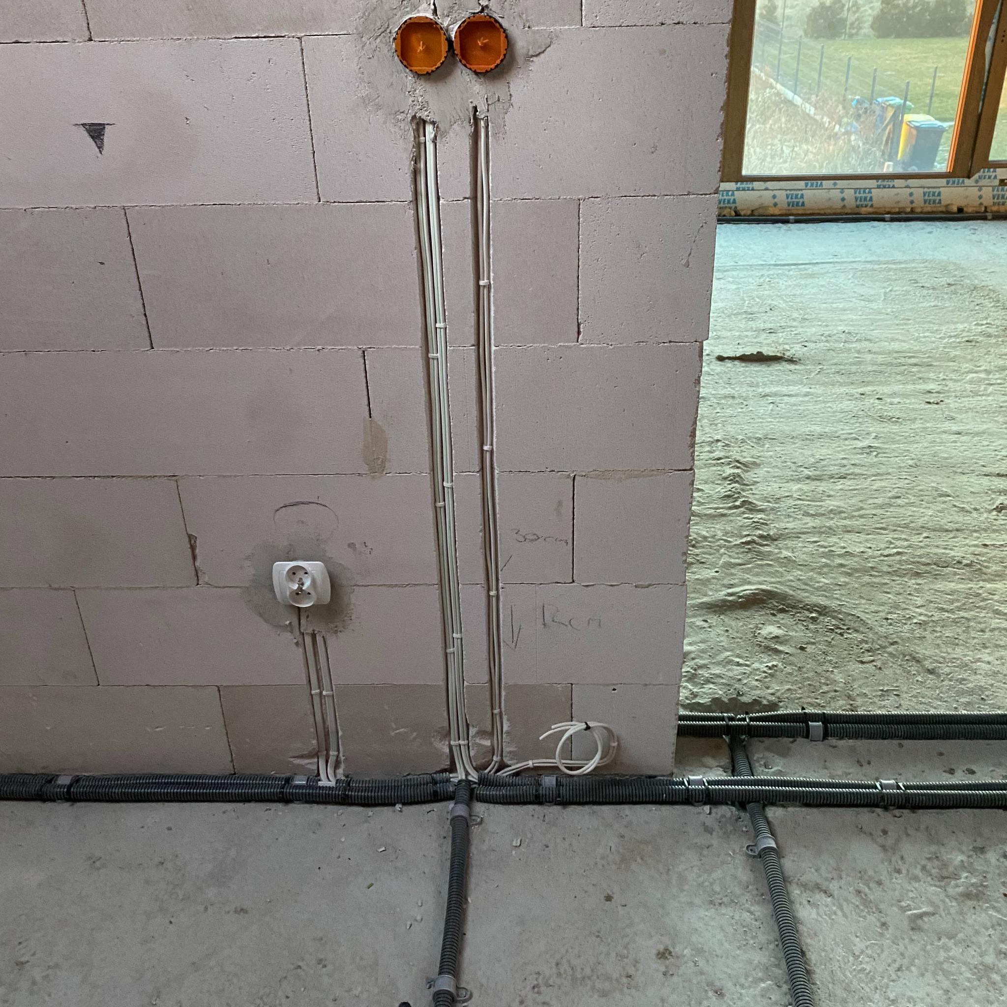 Wewnętrzna instalacja elektryczna w trakcie montażu: przewody w ścianie z betonu komórkowego prowadzone do puszek, gniazdko elektryczne zamontowane, peszle ułożone na podłodze w surowym stanie.