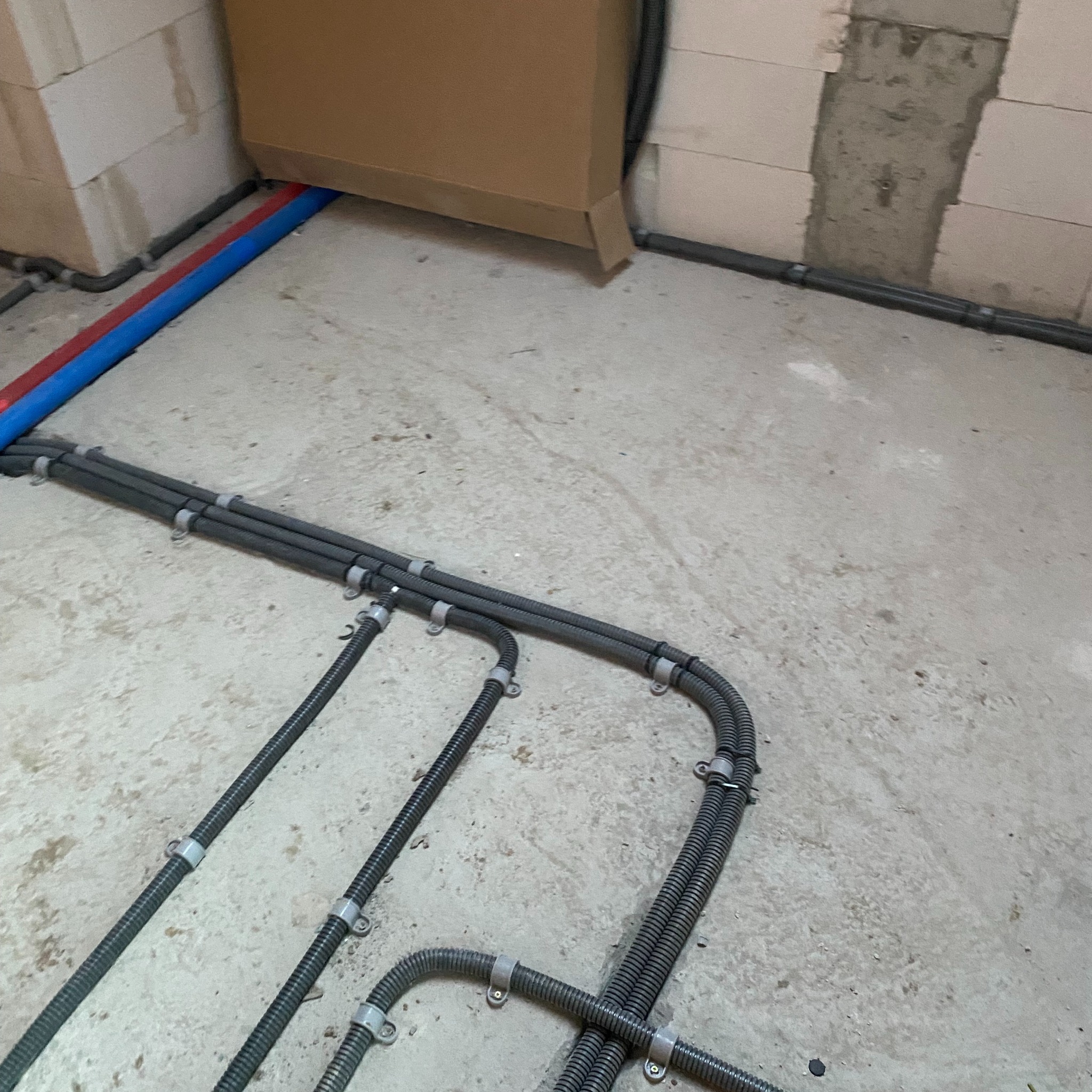 Szare karbowane rury instalacyjne prowadzone po surowym betonowym podłożu, przymocowane metalowymi uchwytami do podłoża i ściany z bloczków. Widoczne rury w kolorze niebieskim i czerwonym.