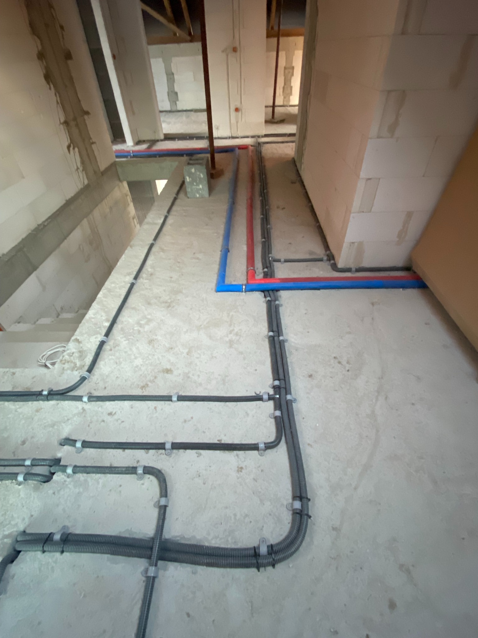 Szare rury karbowane i kolorowe rury (czerwona i niebieska) prowadzone po surowym betonowym podłożu wewnątrz budynku w trakcie budowy, mocowane do ścian klamrami.