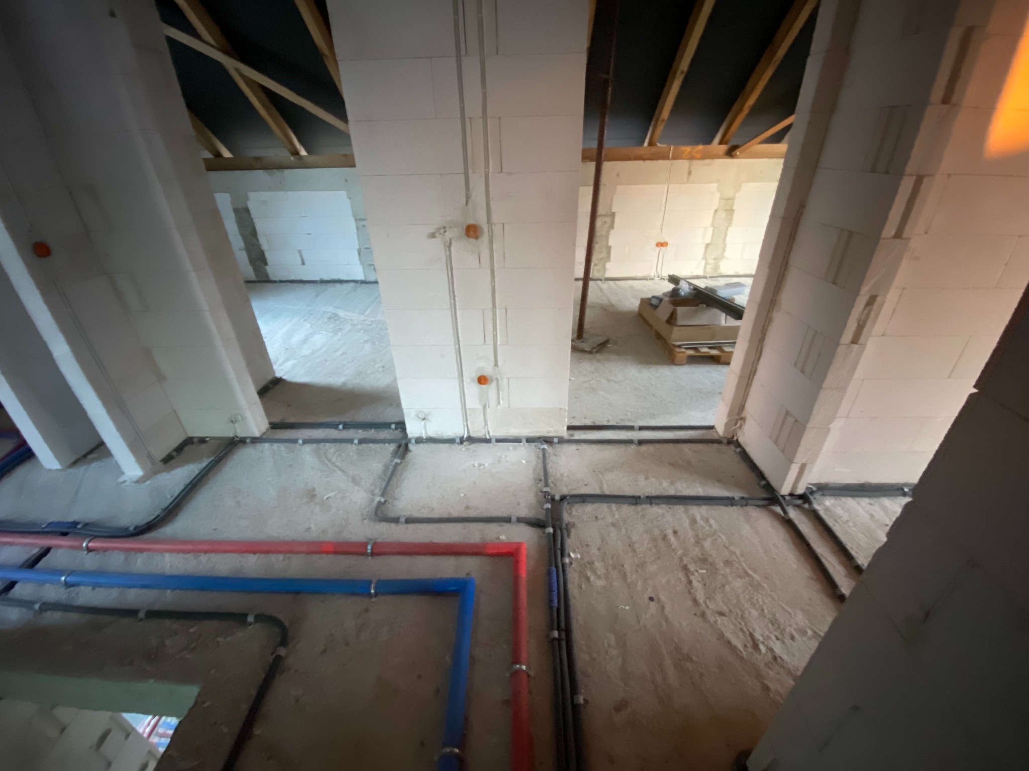 Surowy stan poddasza z widocznymi instalacjami: rurami w kolorze niebieskim i czerwonym, oraz czarnymi kablami elektrycznymi prowadzonymi po podłodze i ścianach z białego bloczka.