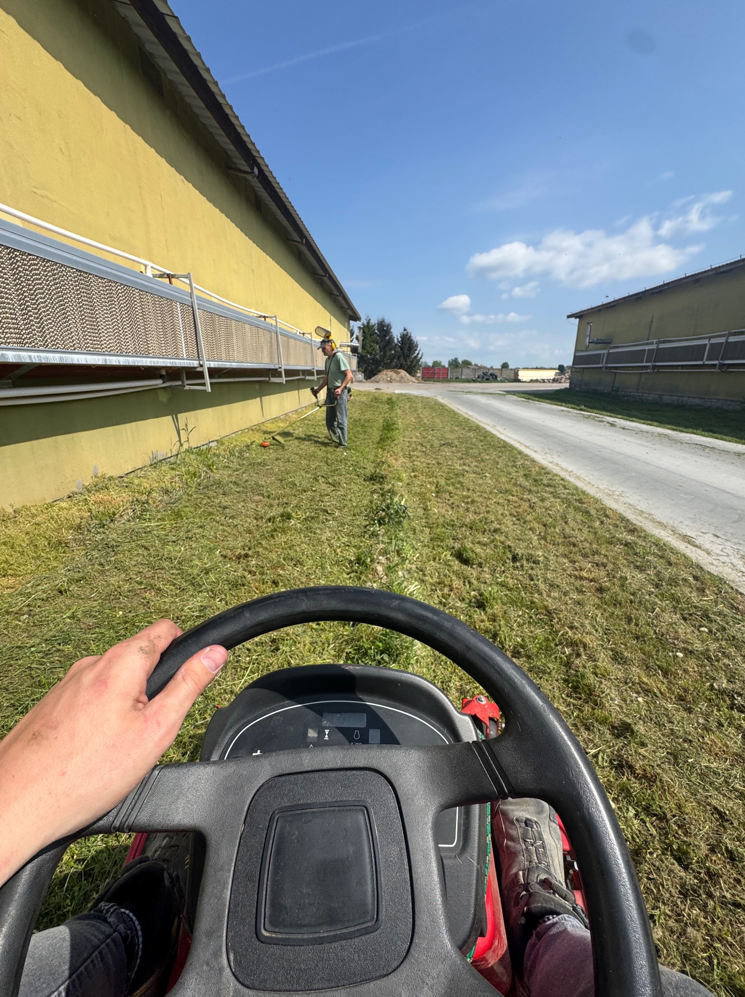 Perspektywa z traktorka koszącego trawę przy budynku. W tle osoba z podkaszarką, świeżo skoszona trawa i fragment drogi. Słoneczny dzień.