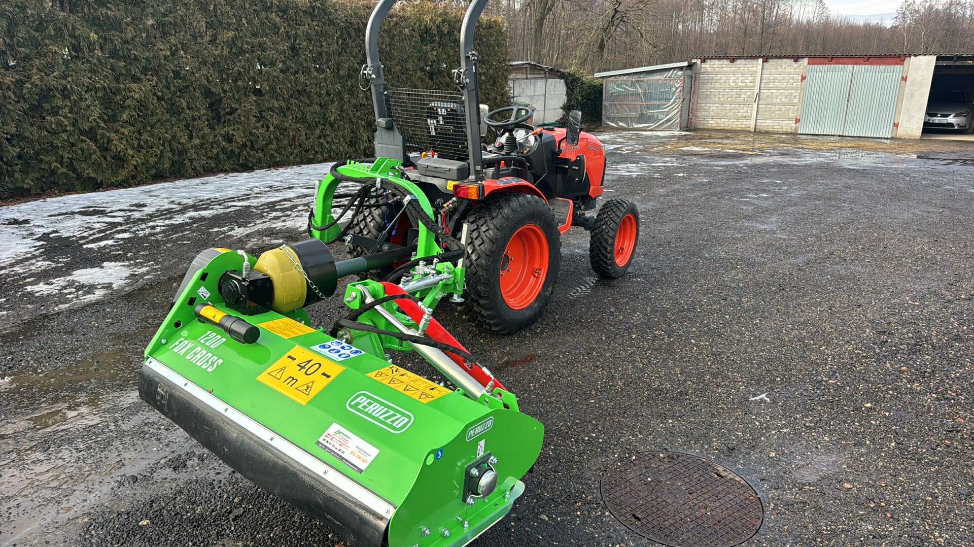 Pomarańczowy traktor z zielonym osprzętem do koszenia trawy Peruzzo Fox Cross 1200 na mokrym podłożu. W tle budynek gospodarczy i żywopłot.