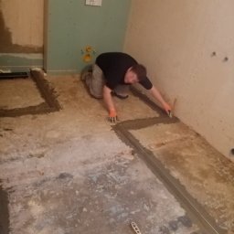 Elephant Bud Sp. Z o. O - Mężczyzna w trakcie przygotowywania podłoża pod posadzkę w łazience, z widoczną konstrukcją podtynkową WC, poziomica laserowa wyznacza linie, kielnia leży na podłodze.