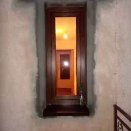 Elephant Bud Sp. Z o. O - Brązowe okno PCV osadzone w surowej, szarej ścianie z widocznymi śladami świeżego tynku wokół ramy, wewnątrz widoczne oświetlone pomieszczenie z drzwiami i butelką na parapecie.