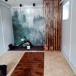 Elephant Bud Sp. Z o. O - Wnętrze w trakcie prac: częściowo pomalowana podłoga na brązowo, drewniana ażurowa ścianka działowa, odkurzacz i wiadro widoczne w tle na tle fototapety z motywem leśnym.