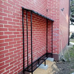 Elephant Bud Sp. Z o. O - Metalowa konstrukcja zadaszenia nad wejściem do budynku z czerwonej cegły, wsparta na betonowych bloczkach, widoczne narzędzia ogrodnicze obok.