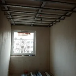 Wnętrze pokoju w trakcie montażu stelażu sufitu podwieszanego z profili metalowych, widoczne profile leżące na podłodze, surowy tynk na ścianach, okno z widokiem na budynek i drzewo.