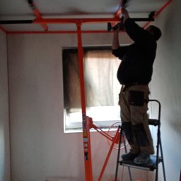 Elephant Bud Sp. Z o. O - Pracownik na drabinie w trakcie montażu płyty kartonowo-gipsowej do sufitu za pomocą podnośnika, widoczne narzędzia i elementy konstrukcyjne.