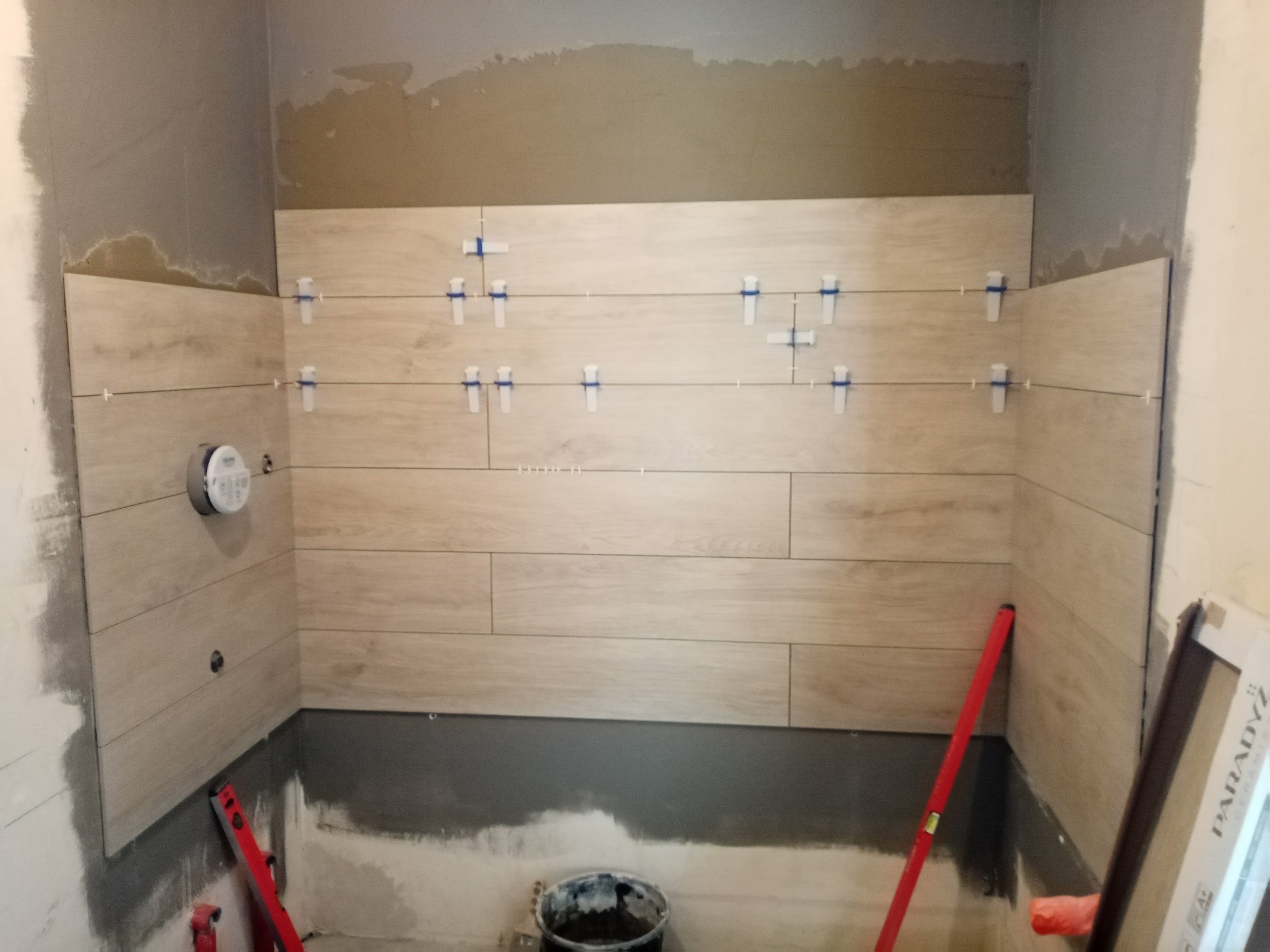 Ściana w trakcie układania płytek imitujących drewno z użyciem krzyżyków dystansowych, widoczny poziomica i przygotowane podłoże w łazience.