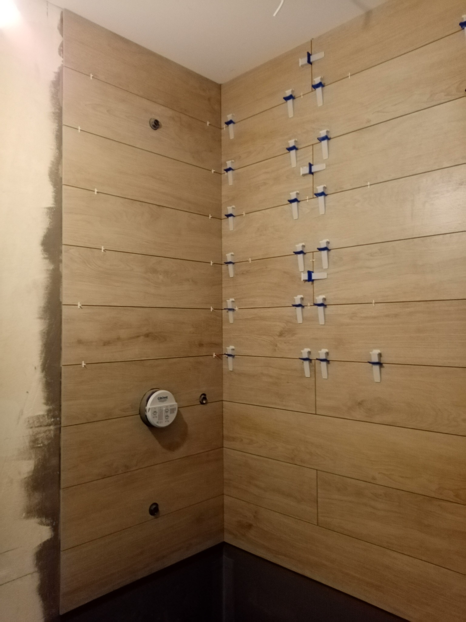 Ściana łazienki w trakcie układania płytek imitujących drewno, widoczne krzyżyki dystansowe i przygotowane otwory na armaturę, surowy fragment ściany obok.