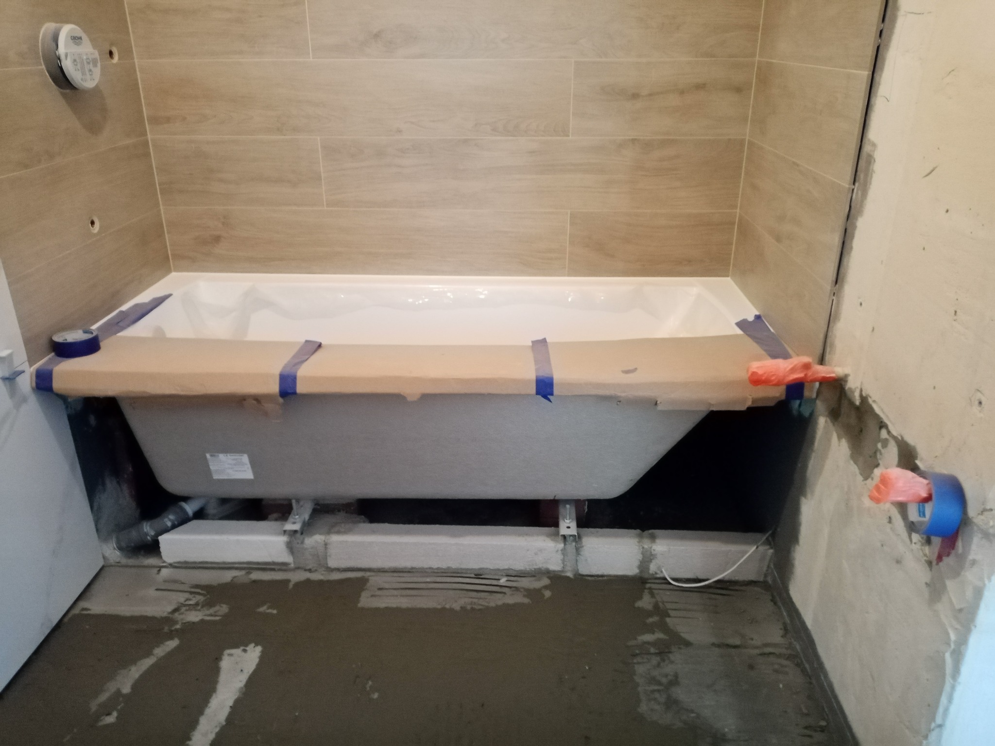 Biała wanna zabezpieczona kartonem i niebieską taśmą, ustawiona na bloczkach z betonu komórkowego w trakcie remontu łazienki z płytkami imitującymi drewno na ścianach.