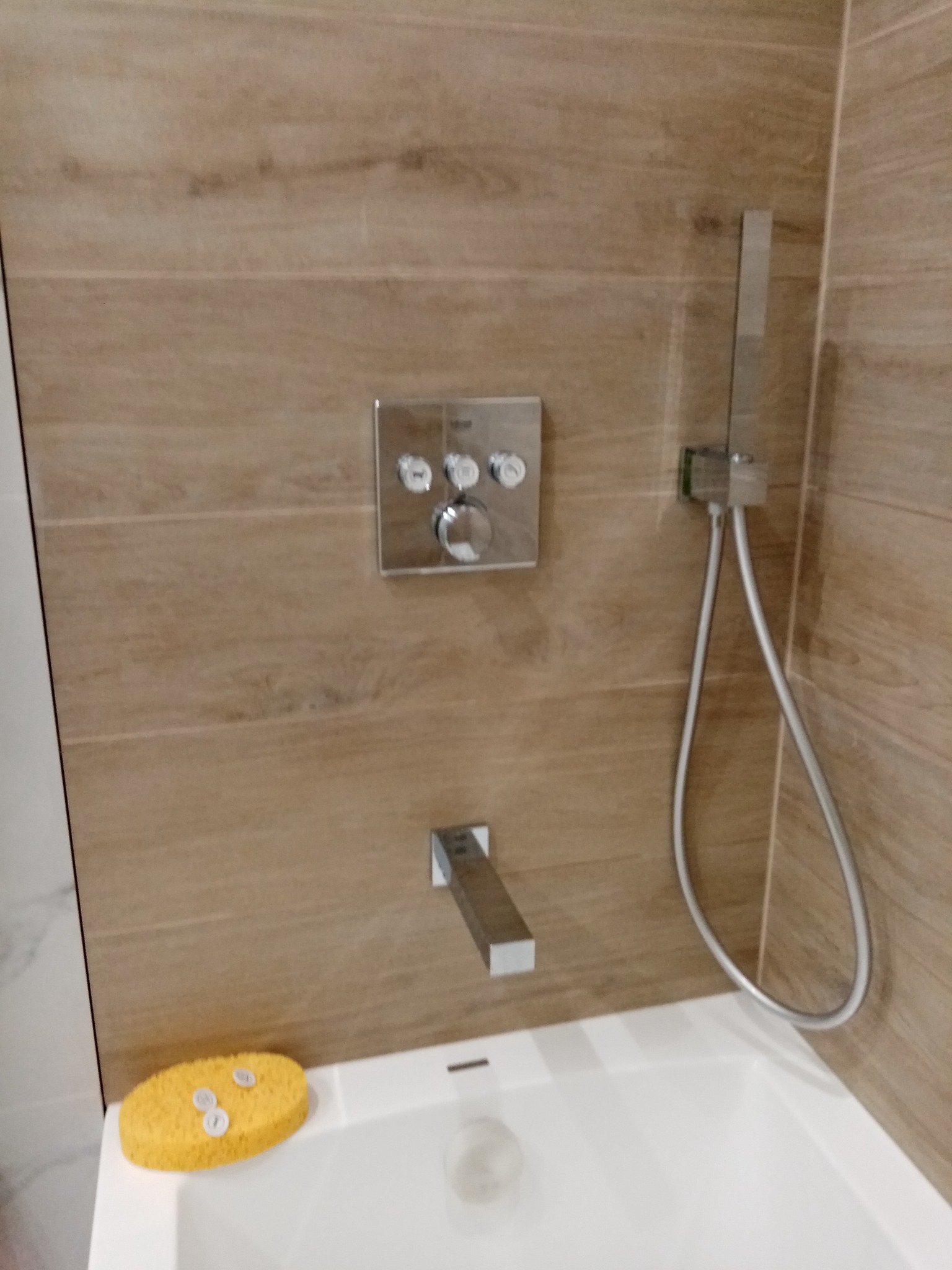 Nowoczesna bateria wannowa z panelem sterującym na ścianie z płytek imitujących drewno, obok słuchawka prysznicowa i żółta gąbka z trzema białymi kapslami na wannie.