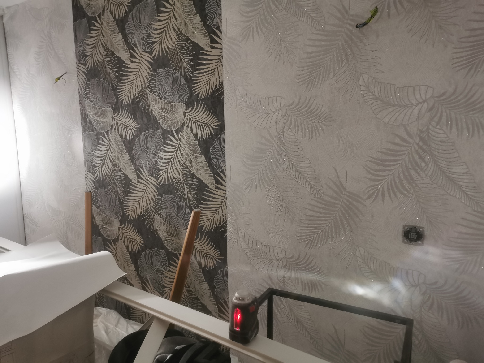 Dwa rodzaje tapet z motywem liści palmowych, jasna i ciemna, na ścianie podczas montażu, widoczny laser krzyżowy i fragment rusztowania.