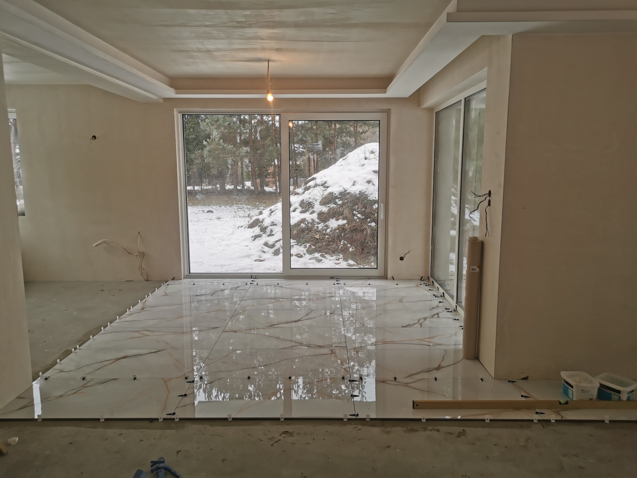 Wnętrze w trakcie układania płytek podłogowych z imitacją marmuru, widoczne krzyżyki dystansowe i poziomica, ściany przygotowane do malowania, duże okno z widokiem na ośnieżony ogród.
