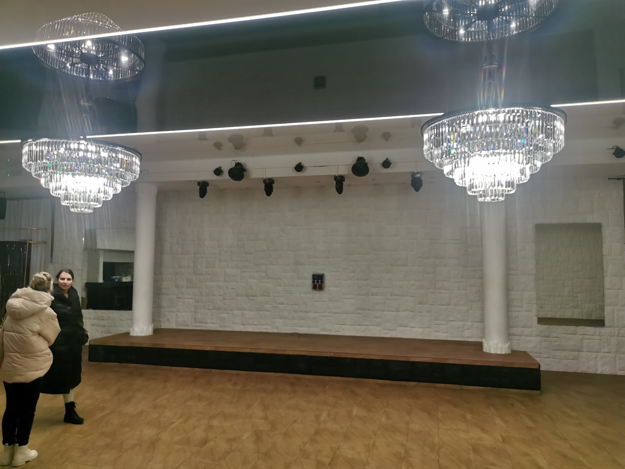 Sala bankietowa z parkietem, białą ścianą z efektem cegły, sceną i kryształowymi żyrandolami. Dwie osoby w kurtkach stoją z boku kadru.