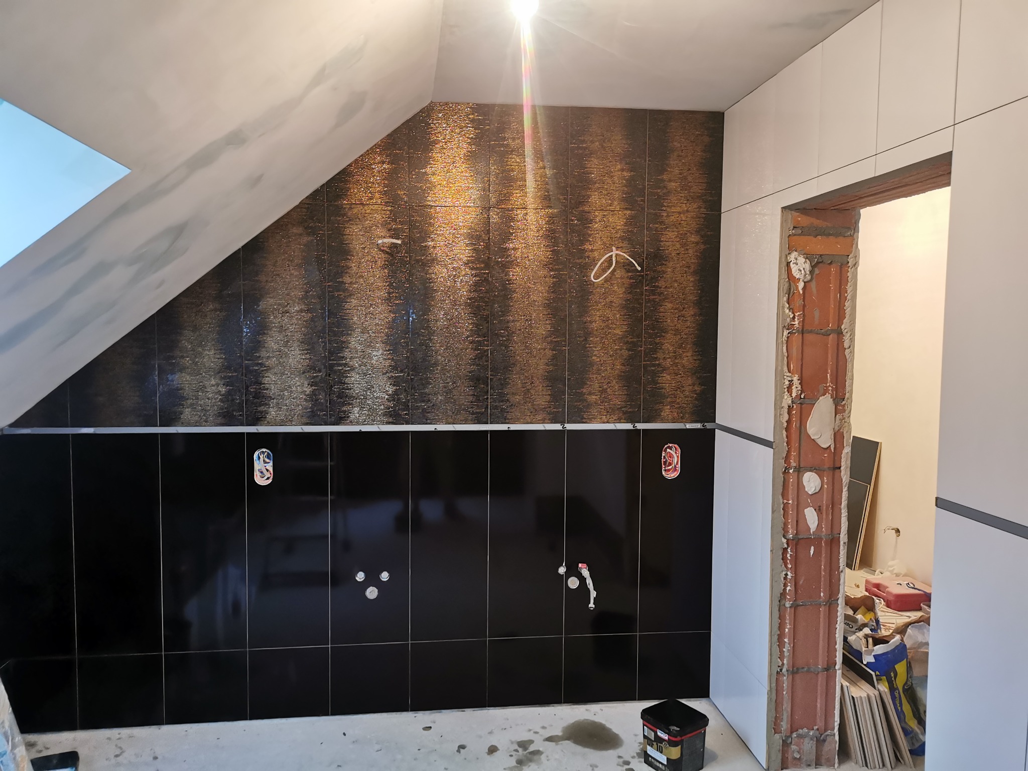 Wnętrze łazienki w trakcie remontu, z częściowo ułożonymi czarnymi i mozaikowymi płytkami, widoczne instalacje hydrauliczne i elektryczne, sufit ze skosem, obudowa ościeżnicy drzwi z cegły...