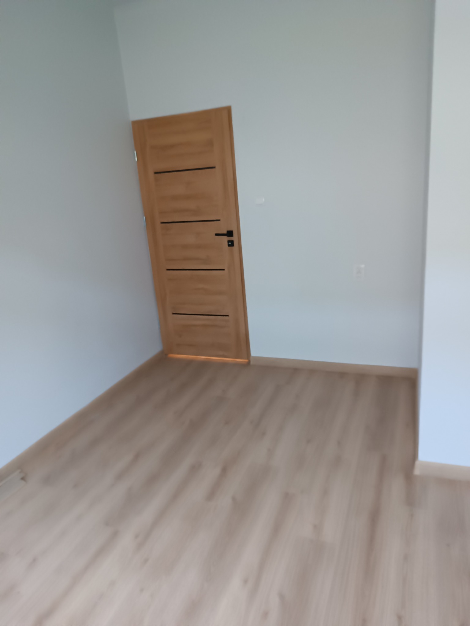 Pusty pokój z nową, jasną podłogą imitującą drewno i świeżo pomalowanymi, jasnoszarymi ścianami; widoczne brązowe drzwi z czarną klamką.