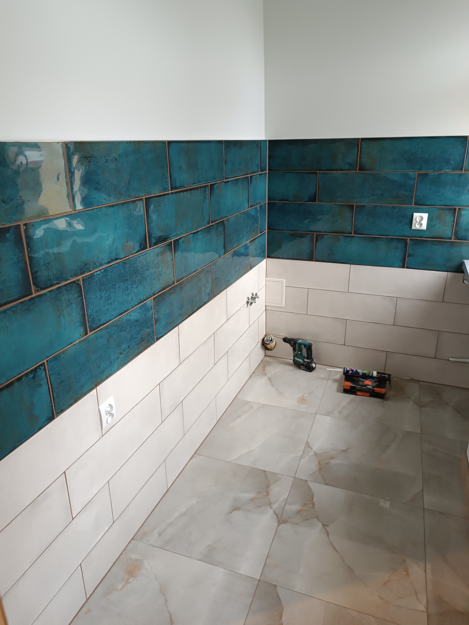 Łazienka w trakcie wykańczania: turkusowe i białe kafelki na ścianach, marmurowe płytki podłogowe, wiertarka akumulatorowa i skrzynka narzędziowa na podłodze, widoczne gniazdka elektryczne...