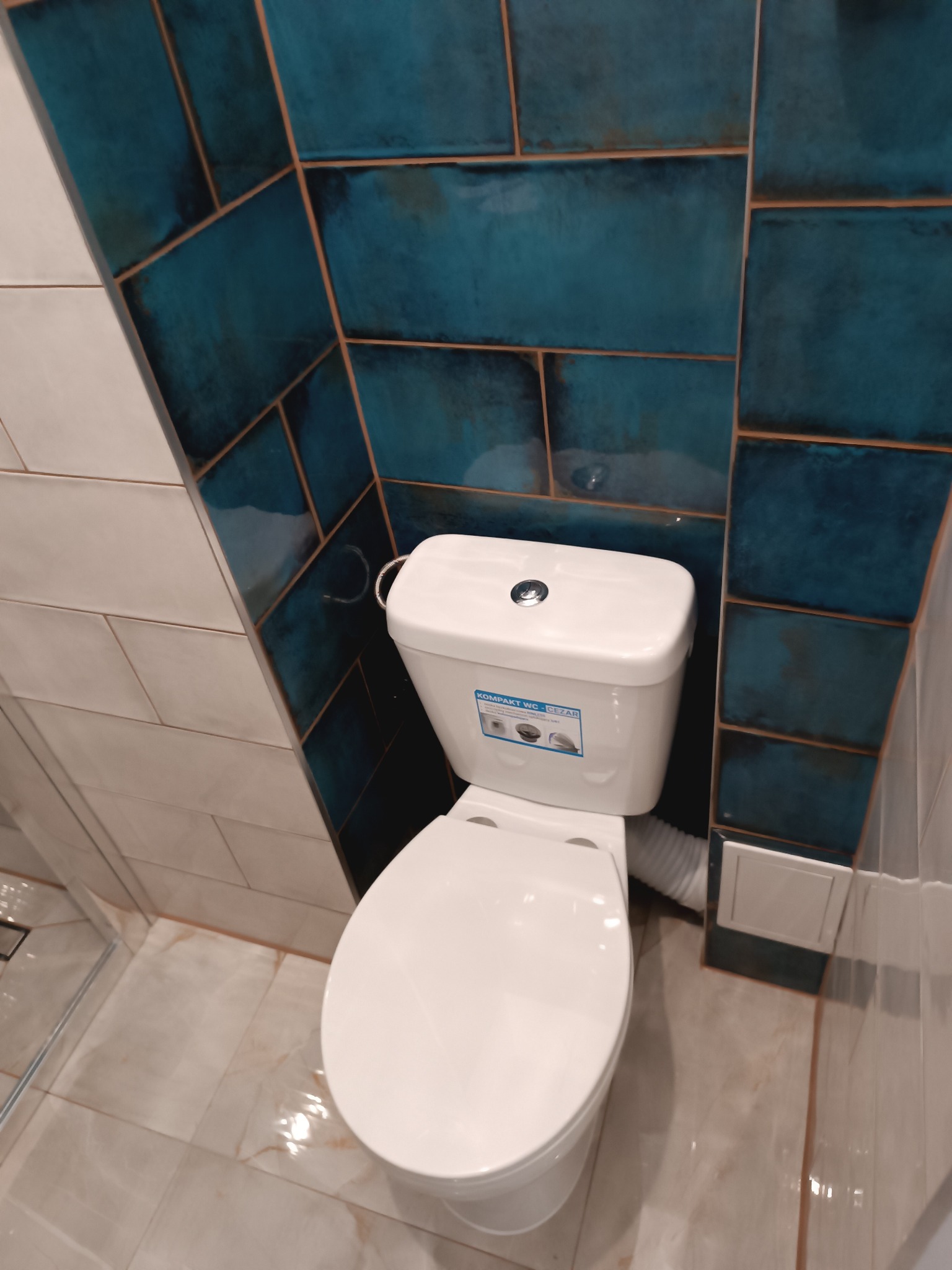 Nowoczesna toaleta kompaktowa z turkusową glazurą na ścianie, widoczna instalacja odpływowa, jasne płytki podłogowe z delikatnym wzorem marmuru, fragment kabiny prysznicowej w tle.