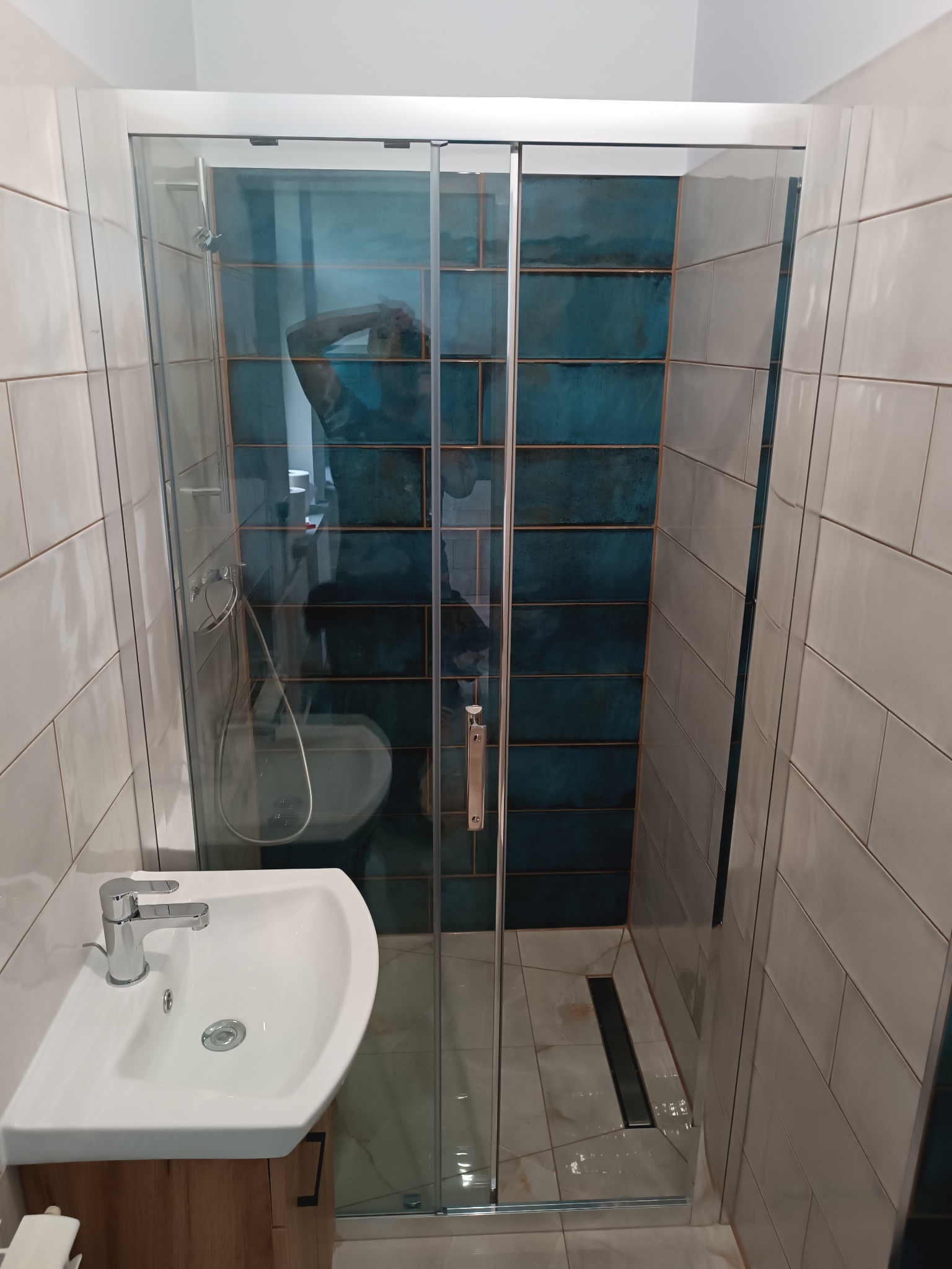 Nowoczesna kabina prysznicowa z przesuwnymi drzwiami, turkusowe i białe płytki na ścianach, biała umywalka z szafką z naturalnego drewna, widoczny odpływ liniowy w podłodze.