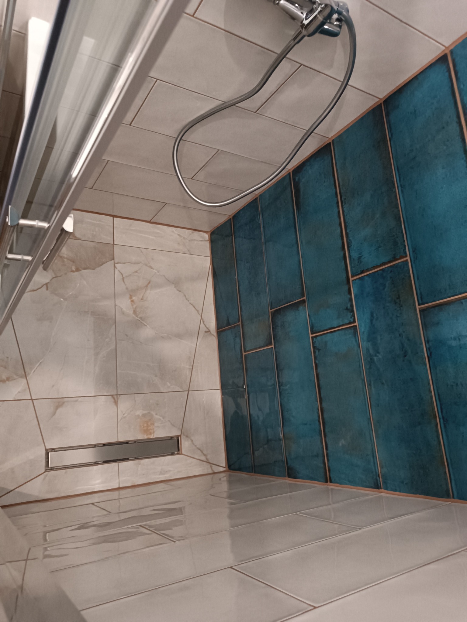 Nowoczesna kabina prysznicowa z odpływem liniowym, wyłożona białymi płytkami imitującymi marmur oraz ścianą z pionowych, turkusowych płytek.