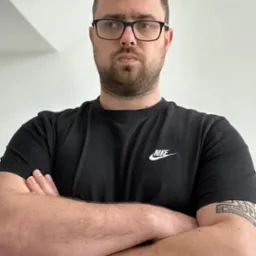 Portret mężczyzny w okularach i czarnej koszulce z logo Nike, skrzyżowane ręce, tatuaż na ramieniu, neutralny wyraz twarzy, białe tło.