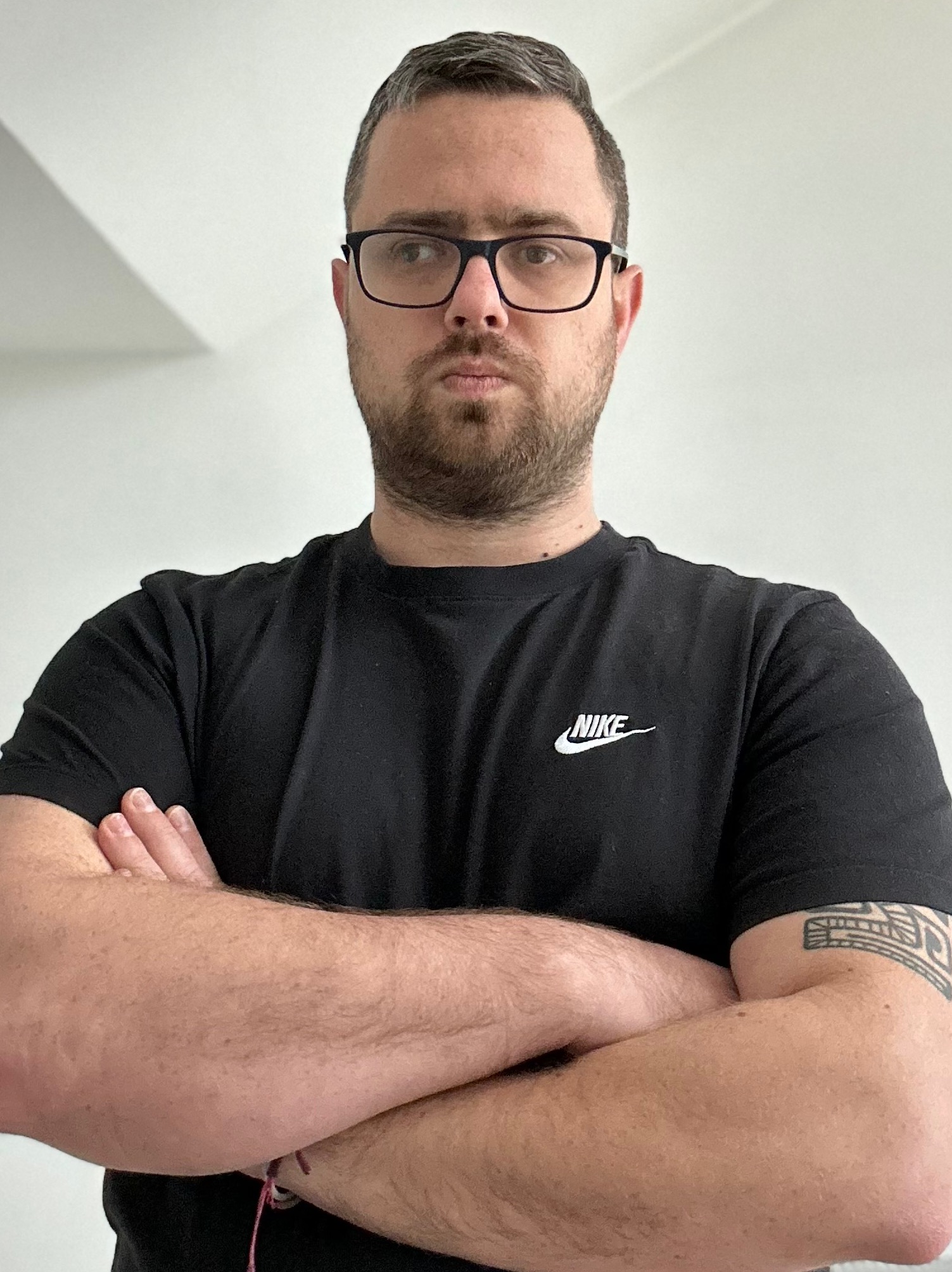 Portret mężczyzny w okularach i czarnej koszulce z logo Nike, skrzyżowane ręce, tatuaż na ramieniu, neutralny wyraz twarzy, białe tło.