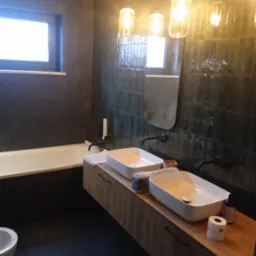 Nowoczesna łazienka z dwiema białymi umywalkami nablatowymi na drewnopodobnej szafce, czarne baterie, ciemne płytki na ścianach, podwieszane lampy z ciepłym światłem i częściowo widoczna czarna wanna.