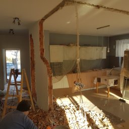 DEKOR M.O. - Pracownik w czapce zbiera gruz po wyburzeniu ściany działowej między pokojem a kuchnią, widoczne odsłonięte cegły i instalacje elektryczne, meble kuchenne zabezpieczone folią.