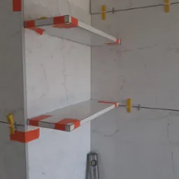 Półki z płytek imitujących marmur w trakcie montażu, zabezpieczone pomarańczową taśmą, widoczne żółte krzyżyki dystansowe i poziomica.