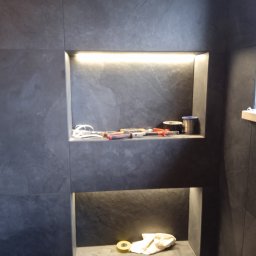 DEKOR M.O. - Półki wnękowe wyłożone szarą płytką w łazience, górna z oświetleniem LED, na której leżą narzędzia i akcesoria glazurnicze, dolna z żółtą puszką i szmatką.