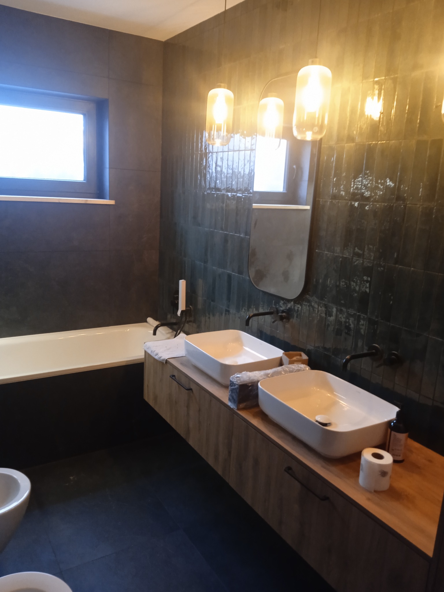 Nowoczesna łazienka z dwiema białymi umywalkami nablatowymi na drewnopodobnej szafce, czarne baterie, ciemne płytki na ścianach, podwieszane lampy z ciepłym światłem i częściowo widoczna czarna wanna.