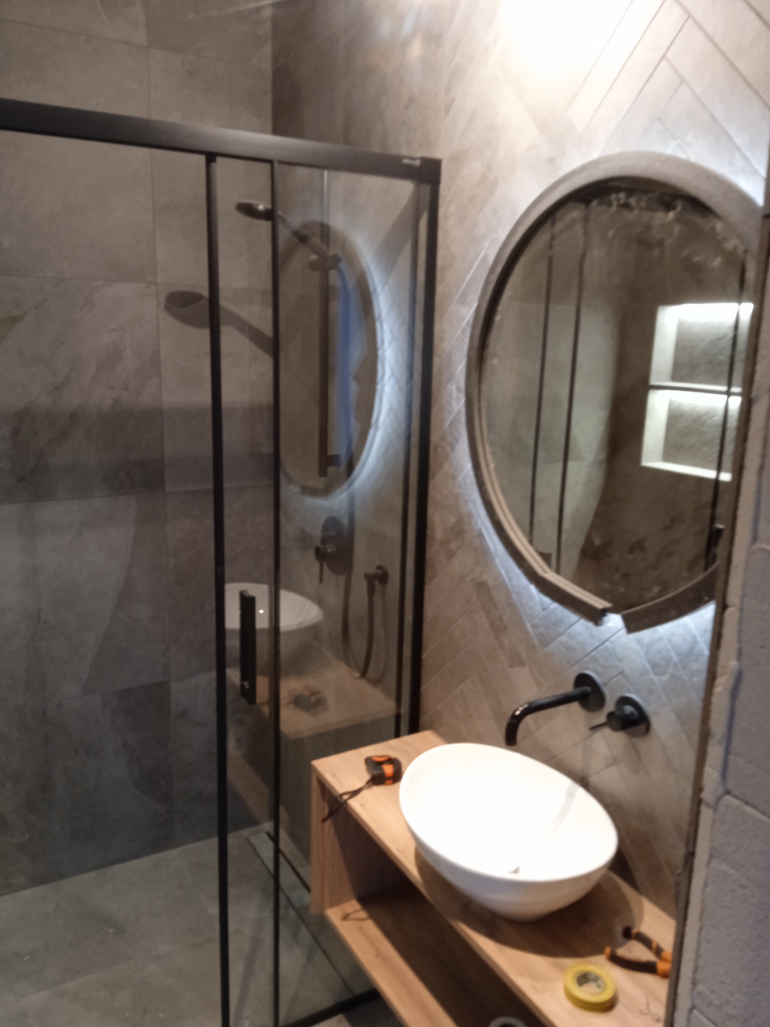 Nowoczesna łazienka z kabiną prysznicową typu walk-in, okrągłym lustrem z podświetleniem LED, białą umywalką nablatową i armaturą w kolorze czarnym; na blacie leżą narzędzia.