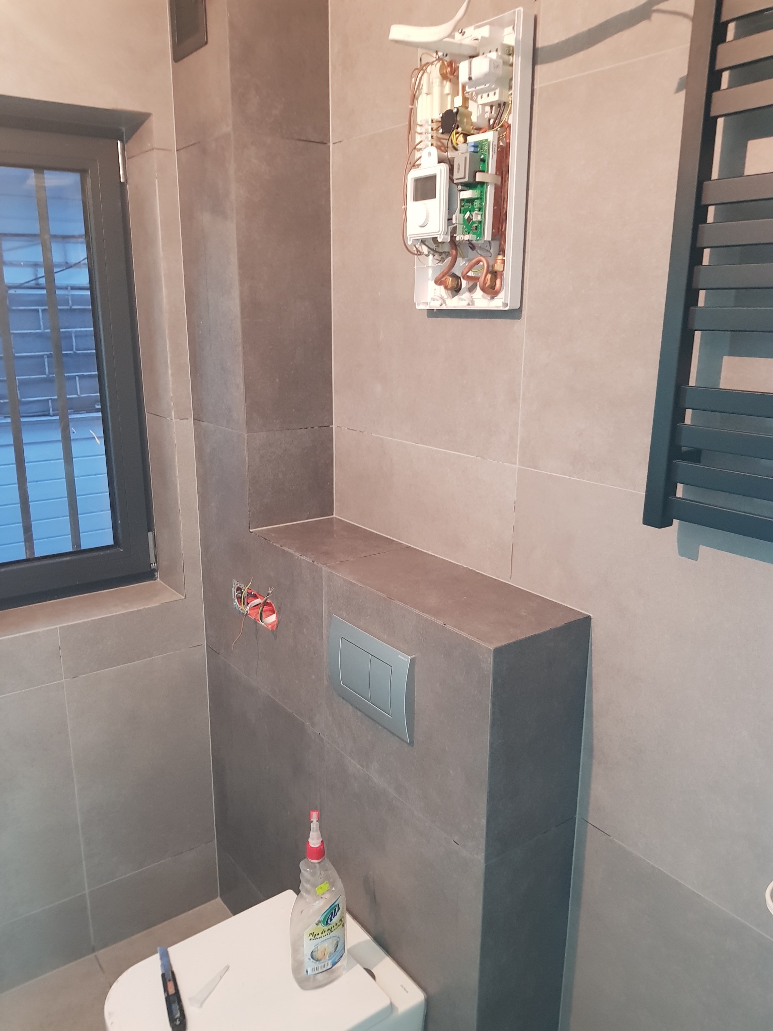 Szare płytki w łazience, zabudowa toalety z przyciskiem spłukującym, otwarty panel sterowania ogrzewaniem wody, grzejnik drabinkowy i butelka płynu do czyszczenia na sedesie.