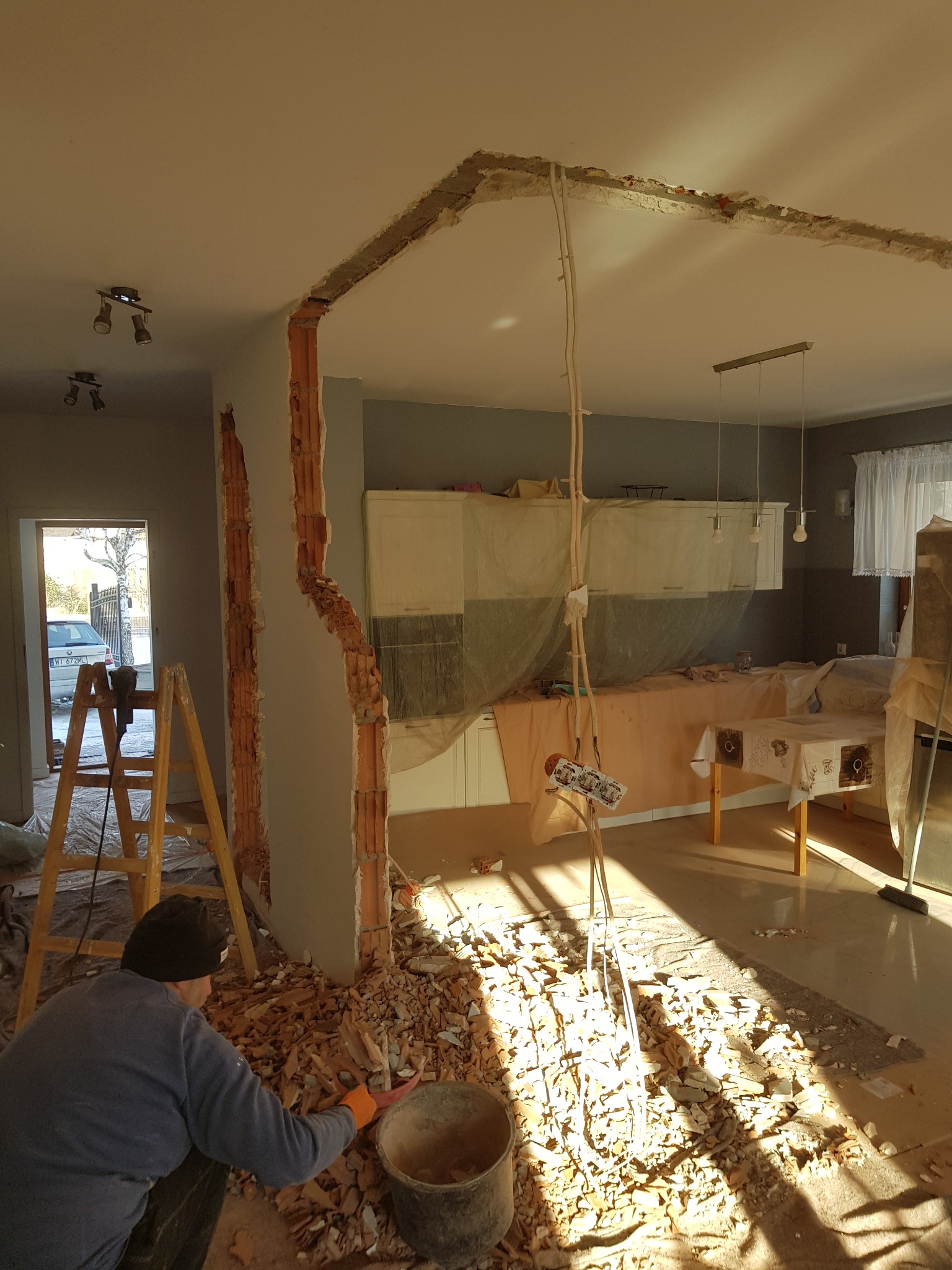 Pracownik w czapce zbiera gruz po wyburzeniu ściany działowej między pokojem a kuchnią, widoczne odsłonięte cegły i instalacje elektryczne, meble kuchenne zabezpieczone folią.