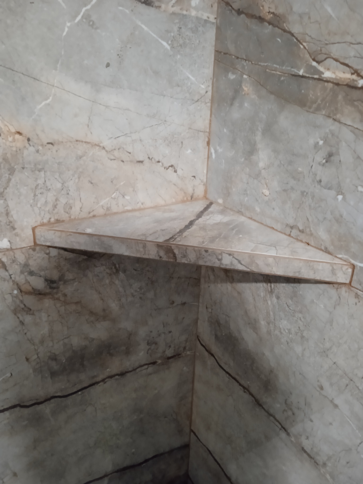 Narożna, trójkątna półka wyłożona marmurowymi płytkami w łazience, dopasowana do okładziny ściennej.