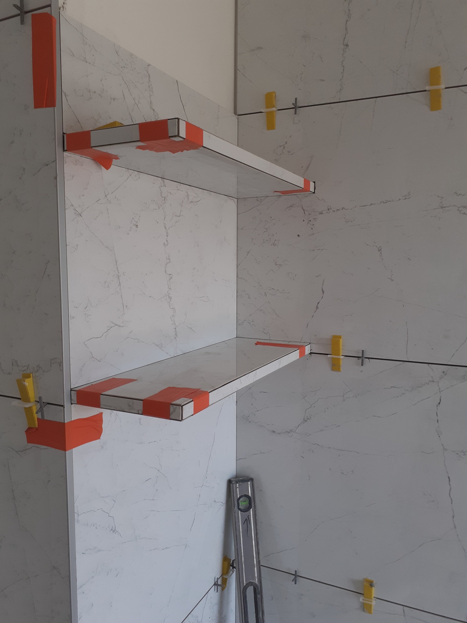 Półki z płytek imitujących marmur w trakcie montażu, zabezpieczone pomarańczową taśmą, widoczne żółte krzyżyki dystansowe i poziomica.