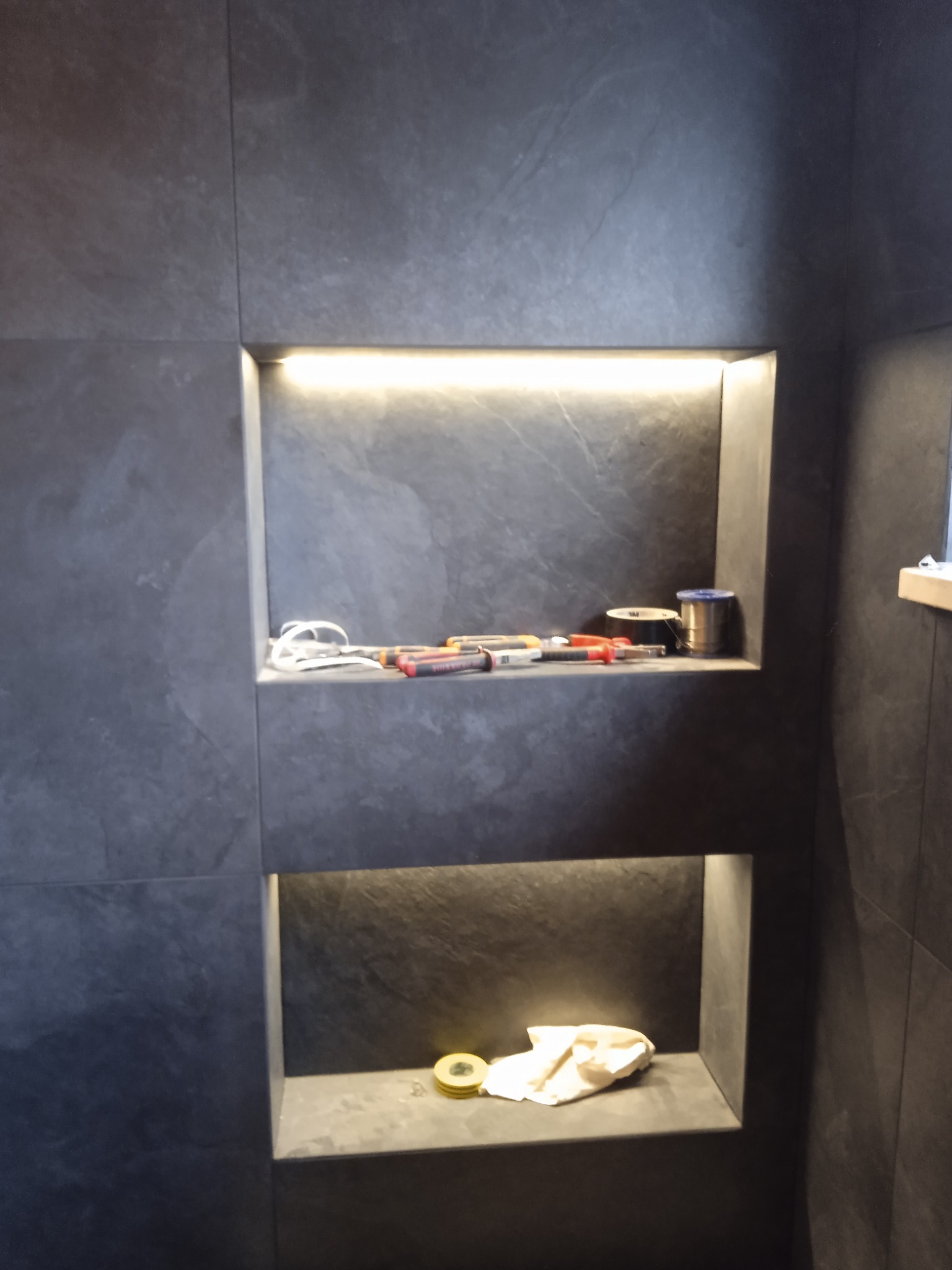 Półki wnękowe wyłożone szarą płytką w łazience, górna z oświetleniem LED, na której leżą narzędzia i akcesoria glazurnicze, dolna z żółtą puszką i szmatką.