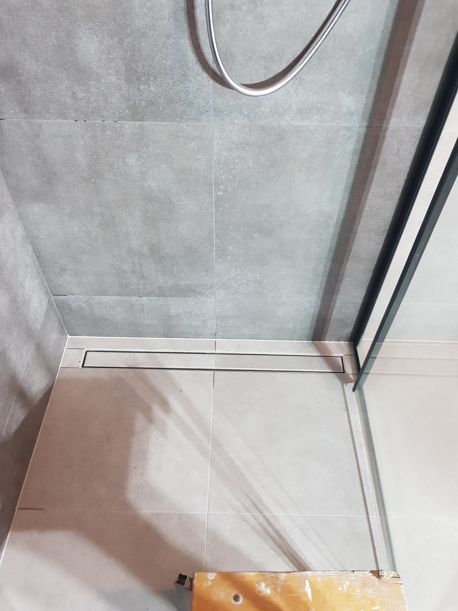 Nowoczesna kabina prysznicowa z szarymi płytkami, odpływem liniowym i fragmentem drabiny malarskiej w rogu.