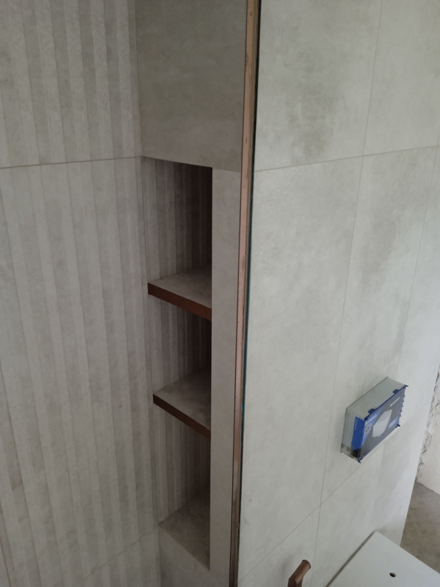 Wykończona wnęka z drewnianymi półkami w łazience, obłożona beżowymi płytkami, widoczna instalacja podtynkowa WC.